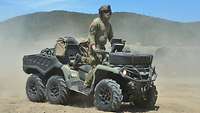 Ein Soldat steht auf einem quadähnlichen Fahrzeug und fährt durchs Gelände, Sand wirbelt auf.