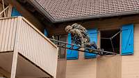 An einer beigen Hauswand klettert ein Soldat über eine schwarze Leiter von Balkon zu Balkon.