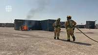 Zwei Personen im Feuerwehranzug laufen mit einem Wasserschlauch auf einen brennenden Container zu.