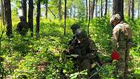Drei deutsche Soldaten hocken im Wald und ein Soldat steht daneben mit einer roten Mappe.