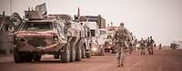 Mehrere Fahrzeuge stehen in einer Reihe auf einer Sandpiste. Daneben stehen und laufen Soldaten.