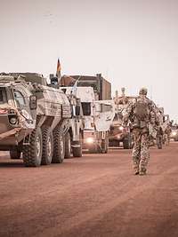 Mehrere Fahrzeuge stehen in einer Reihe auf einer Sandpiste. Daneben stehen und laufen Soldaten.