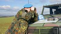 Ein Soldat mit blauem Barett lehnt auf der Motorhaube eines Jeeps und schaut durch ein Fernglas