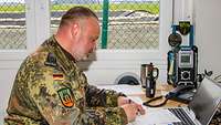 Ein Soldat sitzt in einem Bürocontainer an einem Schreibtisch und füllt eine Liste aus.