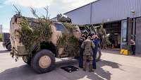 Ein Radpanzer steht vor einer grauen Halle und wird durch Soldaten repariert.