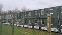 Zu sehen sind aneinander gereihte grüne Container, welche hinter einem Zaun stehen