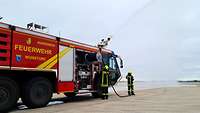 Ein Flugfeldlöschfahrzeug spritzt Wasser über Monitor und 2 Feuerwehrleute über ein Strahlrohr