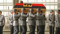 Soldaten tragen einen Sarg, der mit einer Deutschlandflagge umwickelt ist, darauf liegt ein Helm