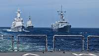 Drei graue Schiffe in Formationsfahrt in See, vorne das Heck eines weiteren Schiffes.