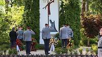 Drei Soldaten, die militärisch grüßen, stehen vor einem Denkmal, vor dem Blumenkränze abgelegt sind.