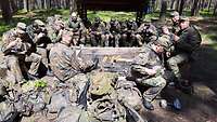 Mehrere Soldaten sitzen auf Bänken, Steinen oder Rücksäcken und essen. 