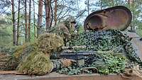 Ein Scharfschütze getarnt auf einem alten Panzer. Daneben kniet sein getarnter Beobachter und funkt.