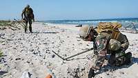 Ein Mann in Flecktarnuniform schaufelt gebückt mit der Hand im Sand eines Strandes.