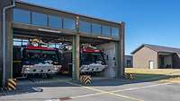 Zwei rote Feuerlöschfahrzeuge stehen in der Garage einer Feuerwache. Die Garagentore sind oben.