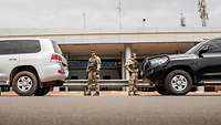 Zwei Soldaten stehen vor einem Flughafen zwischen zwei geparkten Autos.