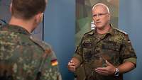 Ein Soldat spricht in einem Studio mit einem Soldaten vor einem Monitor.