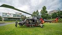 Ein grauer Hubschrauber steht auf einem grünen Rasen vor einem flachen, modernen Gebäude.
