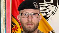 Ein Soldat mit eine schwarzem Barett steht vor einer Truppenfahne.