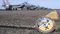 Im Hintergrund sind Düsenjets zu sehen. Im Vordergrund steht ein Patch mit der Aufschrift Air Defender 23