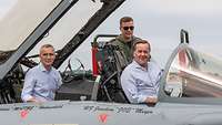 Zwei Männer sitzen im Cockpit. Dahinter steht ein Soldat.