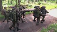 Mehrere Soldaten laufen auf einem Waldpfad und tragen dabei einen Baumstamm.