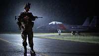 Ein Soldat steht mit Gewehr bei Nacht und Regen auf dem Flugplatz. Hinter ihm steht ein Kampfjet.