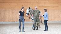 Soldat führt ein Pferd am Halfter und wird links und rechts durch zwei Trainerinnen begleitet
