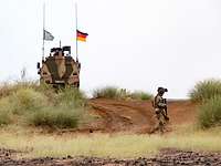 Ein Soldat läuft prüfend im Gelände. Im Hintergrund steht ein Transportfahrzeug vom Typ Dingo.