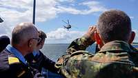 Drei Soldaten schauen auf ein über Wasser fliegenden Hubschrauber.