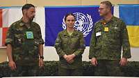 Zwei Soldaten und in der Mitte die Militärbeobachterin stehen nebeneinander vor UN-Flagge.