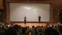 Zwei Soldaten stehen auf der Bühne, hinter ihnen ist eine PowerPoint-Folie zu sehen.
