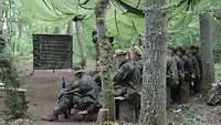 In einem Wald hängt über Bäumen ein alter grüner Fallschirm, darunter sitzen Soldaten auf Bänken.