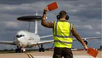 Ein Fluglotse in gelber Warnweste gibt mit zwei roten Signalkellen einem AWACS-Flugzeug Zeichen