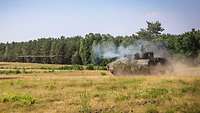 Ein Schützenpanzer steht auf einer umwaldeten Wiese und schießt mit seiner Kanone.