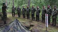 Ein Soldat steht grüßend vor einer Gruppe Soldaten im Wald neben einem Flecktarnzelt.