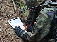 Umgang mit der Sprechtafel Ein Soldat mit Helm und Funkgerät auf dem Rücken schreibt auf einen beigen Zettel.