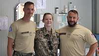 Eine Soldatin flankiert von zwei Soldaten in einem Raum mit Sanitätsmaterial im Hintergrund.