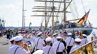 Marinesoldaten in kurzem weißen Diensthemd stehen auf einer Hafenpier und spielen auf Musikinstrumenten.