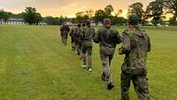 Im Morgengrauen laufen Soldaten auf einer Wiese in einer Reiher hintereinander..