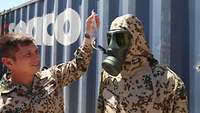 Ein Soldat hält einen durchsichtigen Beutel hoch, ein weiterer Soldat mit Gasmaske trinkt daraus durch einen Schlauch.