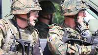Zwei Soldaten mit Helm, Uniform und Gewehren stehen an einem Auto und schauen nach rechts