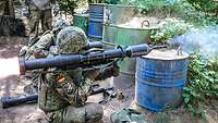 Ein Schützenpanzer hockt hinter einer blauen Tonne und schießt mit einer Panzerfaust.