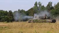 Ein Schützenpanzer steht vor einem Wald auf einer Wiese und schießt mit seiner Kanone.