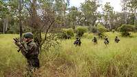 Mehrere Soldaten laufen mit Abstand zueinander durch hohes Gras.