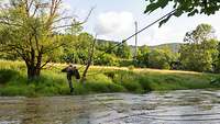 Eine Person in Uniform liegt auf einem Seil. Sie zieht sich daran über einen Fluss.