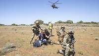 Zwei Soldaten überprüfen am Boden einen Zivilisten. Im Hintergrund fliegt ein Hubschrauber. 