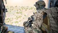 Ein Soldat sitzt an der offenen Tür eines fliegenden Hubschraubers. Im Hintergrund trockene Landschaft.