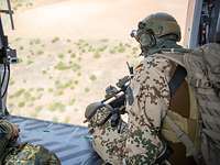 Ein Soldat sitzt an der offenen Tür eines fliegenden Hubschraubers. Im Hintergrund trockene Landschaft.