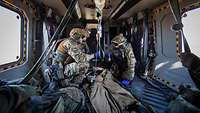 Drei Soldaten knien im Inneren eines Hubschraubers um eine Person herum, die zwischen ihnen liegt und eine Infusion erhält. 
