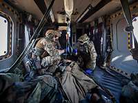 Drei Soldaten knien im Inneren eines Hubschraubers um eine Person herum, die zwischen ihnen liegt und eine Infusion erhält. 